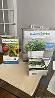 Photo showing boxed AeroGarden Harvest indoor garden with two additional boxed 6 pod kits labeled Grow Anything and Gourmet Herb on table.