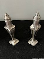 Front view of the pair of Art Deco style chrome salt and pepper shakers on a black surface showing detailed geometric star-shaped bases and ridged tops.