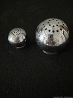 Photo of two small, round, shiny silver salt and pepper shakers on a black surface showing their top dispensing holes and reflective finish.