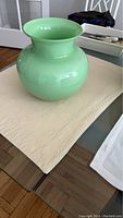 Light green glossy glass vase with rounded body and wide flared mouth shown on a table with a cloth under it.