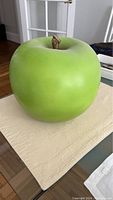 Front and top view of a large green apple decoration placed on a beige cloth on a glass table, showing smooth bright green surface and a small brown stem.