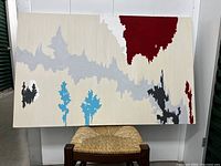 Photo showing modern abstract oil painting on canvas with jagged shapes in red, grey, blue, black, and white on neutral background, positioned resting on woven seat stool.