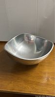 Front angle view of Nambe tri-corner silver alloy bowl on wooden surface, showing overall shape and reflective surface.
