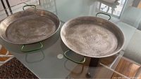 Two round metal paella pans on glass table showing top surfaces and green side handles