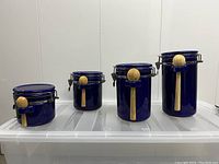 Four cobalt blue ceramic airtight canisters with metal locking lids and attached wooden spoons arranged left to right from shortest to tallest.