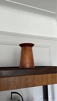 Photo showing wooden vase or container on a wooden shelf against a white wall. The item is tall with a slightly flared lip at the top, natural wood tone.