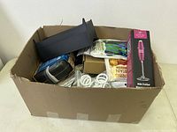 Box containing assorted household kitchen items, including a boxed milk frother, black rack, spiral coils, and packaged goods.