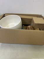 Box viewed from above showing large white ceramic bowl, tiki mug, blue bottle, several cardboard boxes and packaged cards.