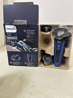 Electric shaver with three rotary blades, charging cable, and protective covers in molded cardboard packaging beside original Philips 5000 Series box.