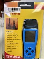 Close-up photo of blue handheld EMF tester in yellow and clear retail packaging showing printed features and specifications.