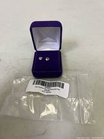 Pair of sterling silver paw print stud earrings in a purple jewelry box with white satin lining, placed on a surface with the original plastic packaging in front showing the product label.
