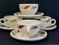 Set of four white ceramic tea cups, each on matching saucers. All have colored leaf designs around rim. Grouped stacked against black background.