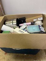 Large cardboard box filled with various items including travel mugs, storage containers, boxes, and personal/fishing accessories.