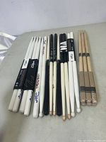 Photo showing 12 pairs of drum sticks laid out on table, various brands and colors including white, black, and natural wood finishes.