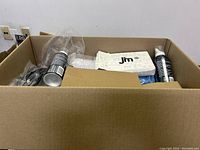 Top view of large heavy box filled with various craft, home, and tool accessory items including black spray cans, white labeled boxes, and assorted packaged goods.