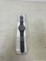 Swatch watch inside its clear plastic case, showing the front view of the watch with black strap and dial.