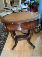 Front and side view of an oval antique drum table with worn leather top, wooden drawer, and metal claw feet with wheels.