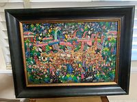 Framed oil painting showing a colorful and detailed Balinese scene with many figures and decorative elements. The frame is black with a gold inner edge.