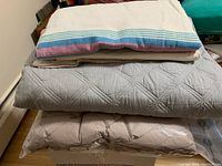 Stack of blankets showing grey quilted blanket and brown-grey duvet-type blanket at bottom in plastic