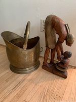 Photo shows large hammered brass pot with curved handle beside a wooden statue of a woman bending over a child in a basin, on wood floor against white wall.
