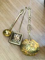 Three antique brass utensils including a cream skimmer spoon, bed warmer, and soup spoon displayed on a wooden floor, showing perforations and decorative handle designs.