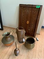 Photo of five items: wooden tray leaning against wall with visible inlay and brass handles; in front three brass/copper vessels with porcelain handles and one copper vessel; one item is a brass oil lamp.