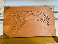 Top view of rectangular wooden carving with engraved First Nations bird design and brass corners