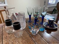Photo showing 2 clear hand-blown glass carafes with black ribbed tops, 2 heavy crystal-like glasses, 4 blue stemmed glasses, decorative blue and gold teacup with saucer, and butterfly glass teacup with saucer.