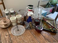 Photo showing a variety of household items including ceramic bottles, wooden stands, cobalt jar, bowling ball shakers, seahorse napkin holders, antique plate, chopsticks bundled, and other small items on wooden table.