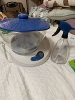 Lotus Sanitizing System with clear plastic bowl, blue perforated lid, white base with digital control panel, and spray bottle on bed