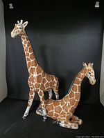 Two glazed ceramic giraffe sculptures against black background, one standing tall and one reclining.