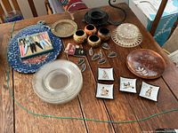 Photo of the entire lot showing woven pot holders, cast iron candle holder, mini smoky glass dishes, brass plate, small vases, wooden bowl, glass plates, and decorative square plates.