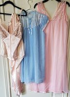Three vintage sleepwear pieces on hangers: a satin pink two-piece, a blue sheer nightgown with lace, and a pink sheer nightgown with lace neckline.