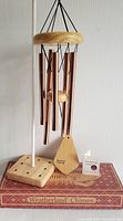 Full view of Weatherland bronze wind chimes hanging next to their original box and a stand (not included). The chimes have an oak wood top ring and several bronze tubes.