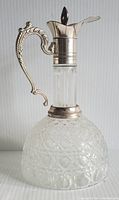 Front angle view of vintage pressed glass decanter with silver plate top and ornate handle showing bell bottom and fluted neck.
