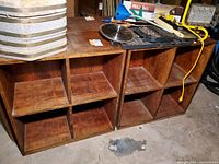 Two wooden shelving units with four compartments each shown side by side in a basement setting, surfaces with scuffs and scratches.