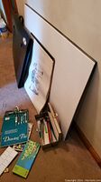 Photo shows multiple foam core boards leaning against wall along with a black portfolio case and a large sheet of paper with sketch. On floor are several art supplies including a drawing pad, paint brushes, and paint containers.