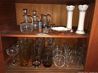 Shelf with Lenox candle holders, oil and vinegar bottles, drinking glasses, shot glasses, salt and pepper shakers, and a decorative glass dish.