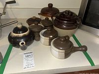 Seven pieces of brownware ceramic pots and crocks with lids displayed on a countertop including a large two handled pot, smaller lidded pots, and teapot.
