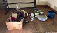 Wide view showing all items including box of Crate & Barrel ornaments, clear glass hurricane candle holder, metal menorah, kerosene lanterns, and various ceramic plant pots.