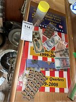 Drawer with sealed US state quarters collector map, several banknotes, sheets of postage stamps, and coins in a yellow capped plastic tube.