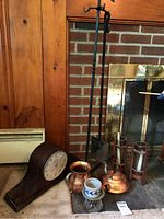 Full lot image showing the clock, copper pitcher, copper kettle, candle holders, and fireplace tool stand in front of brick fireplace and wooden paneling.