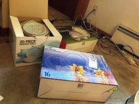 Three boxed sets including Crisa clear glassware set on top, stoneware 20 piece set box on left, and Siestaware porcelain set box in back with floral plates.