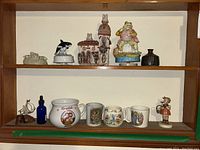 Overview of shelf with assorted collectible figures, mugs, folk art items including Bunnykins, Campbell Soup mugs, Hummel figurine, spun art cottage, ship figure, and toad music box.