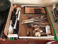 Photo showing the set of cutlery knives with black handles and a variety of spoons inside a cardboard box.
