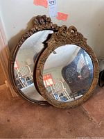 Two round mirrors leaning against a wall on carpet, details of the brass-toned enameled wood frames visible. Larger mirror has smooth ruffled edges.