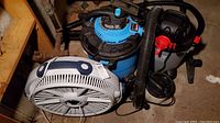 Photo showing two shop vacuums and one table fan positioned closely. Visible hoses and attachments attached to the shop vacuums. Fan is white with blue accents.