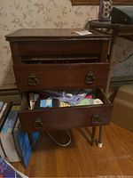 Side angle view of a dark wood side table with two drawers open and filled with sewing notions such as thread, tape, and needles.