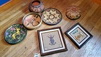 Eight art and pottery items including five bowls, one woven basket, one carved colorful plate, two framed Native American sand art paintings. All shown on wooden floor near blue and brown walls.