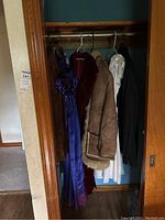 Full view of hanging vintage clothing items inside a closet including purple satin gown, burgundy velvet gown, brown Sherpa jacket, white dress, black tuxedo pants